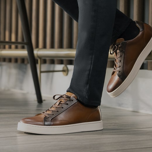 The image features a model leaning against a brick wall while wearing gray pants and brown and blue Men’s boots. Shop the Men’s Dress Boots category now.