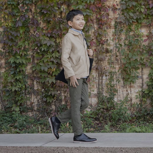 The image features a kid wearing a blue blazer, khaki pants, and brown Kids shoes while standing on a front stoop. Shop the Kids Shoes category now. 